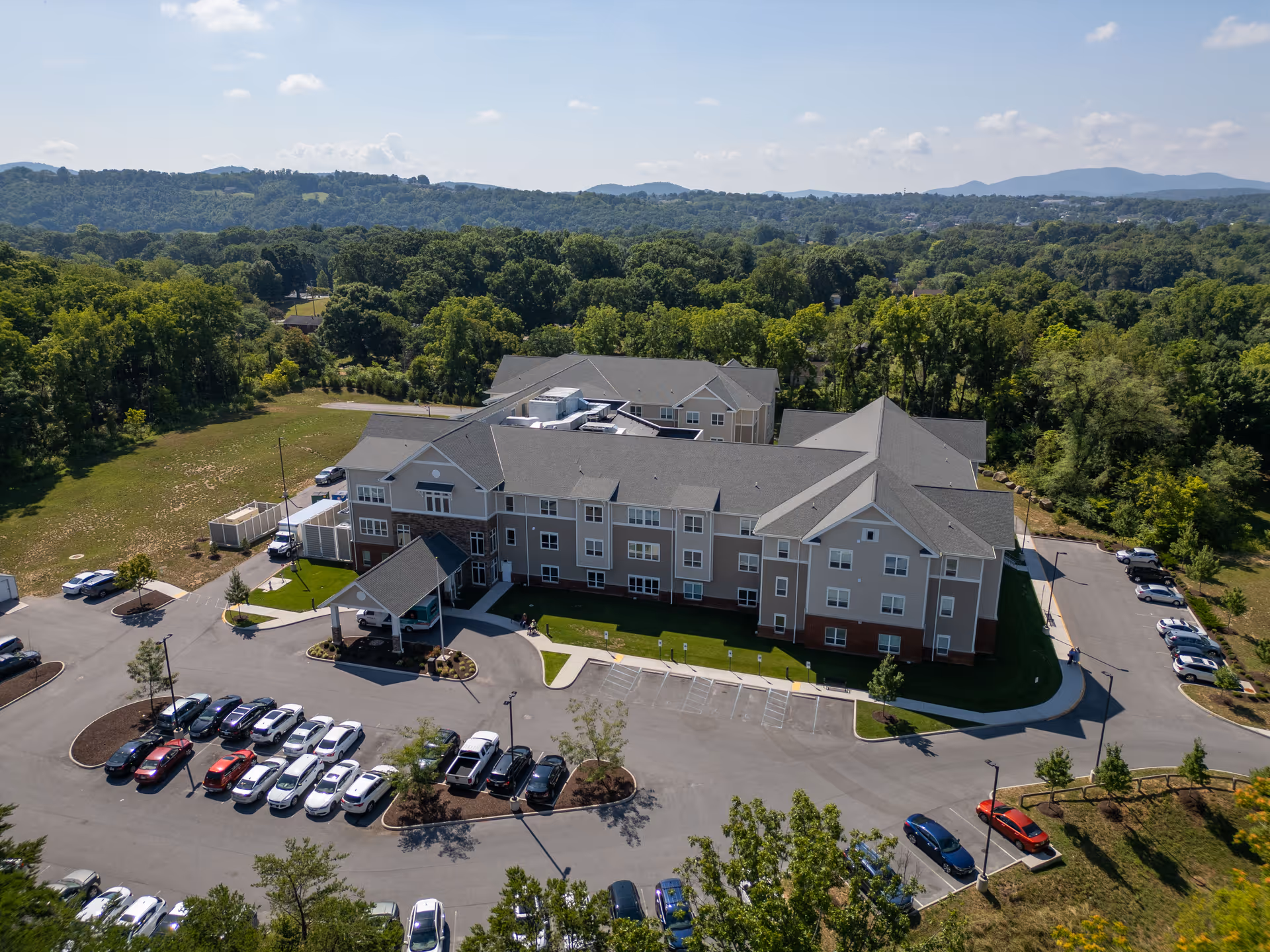 Aerial view of a three-story senior care building with a covered entrance, surrounding parking lot, trees and distant hills.