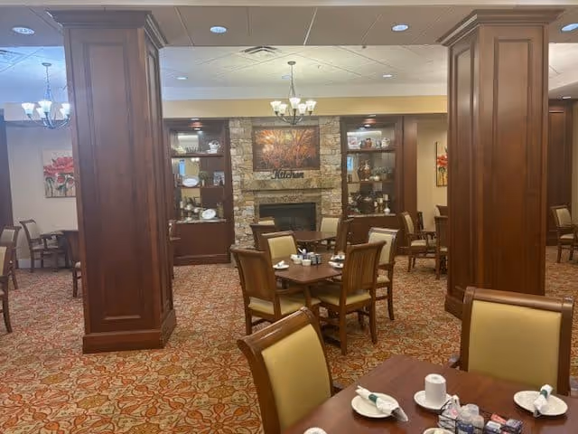 A dining room in a senior living facility featuring multiple wooden tables and chairs with beige cushions. The room has a patterned carpet, wooden pillars, and a stone fireplace with decorative shelves on either side. There are chandeliers hanging from the ceiling and framed artwork on the walls.