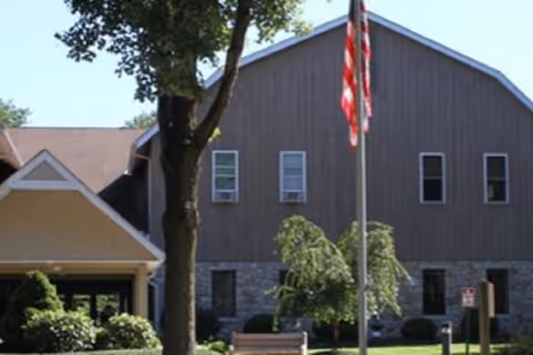 Exterior view of a senior living facility building with a stone and wood facade, an American flag on a flagpole in front, a tree, and some bushes and landscaping.