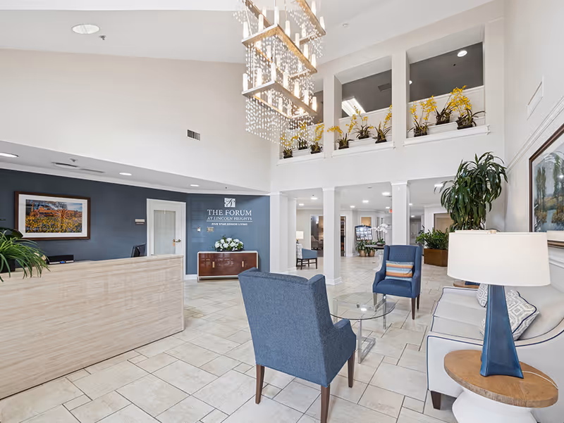 Bright and spacious senior living facility lobby with a modern reception desk, blue armchairs, a glass coffee table, a white sofa, and decorative plants. A large chandelier hangs from the ceiling, and a sign on the wall reads 'The Forum at Lincoln Heights Five Star Senior Living.'