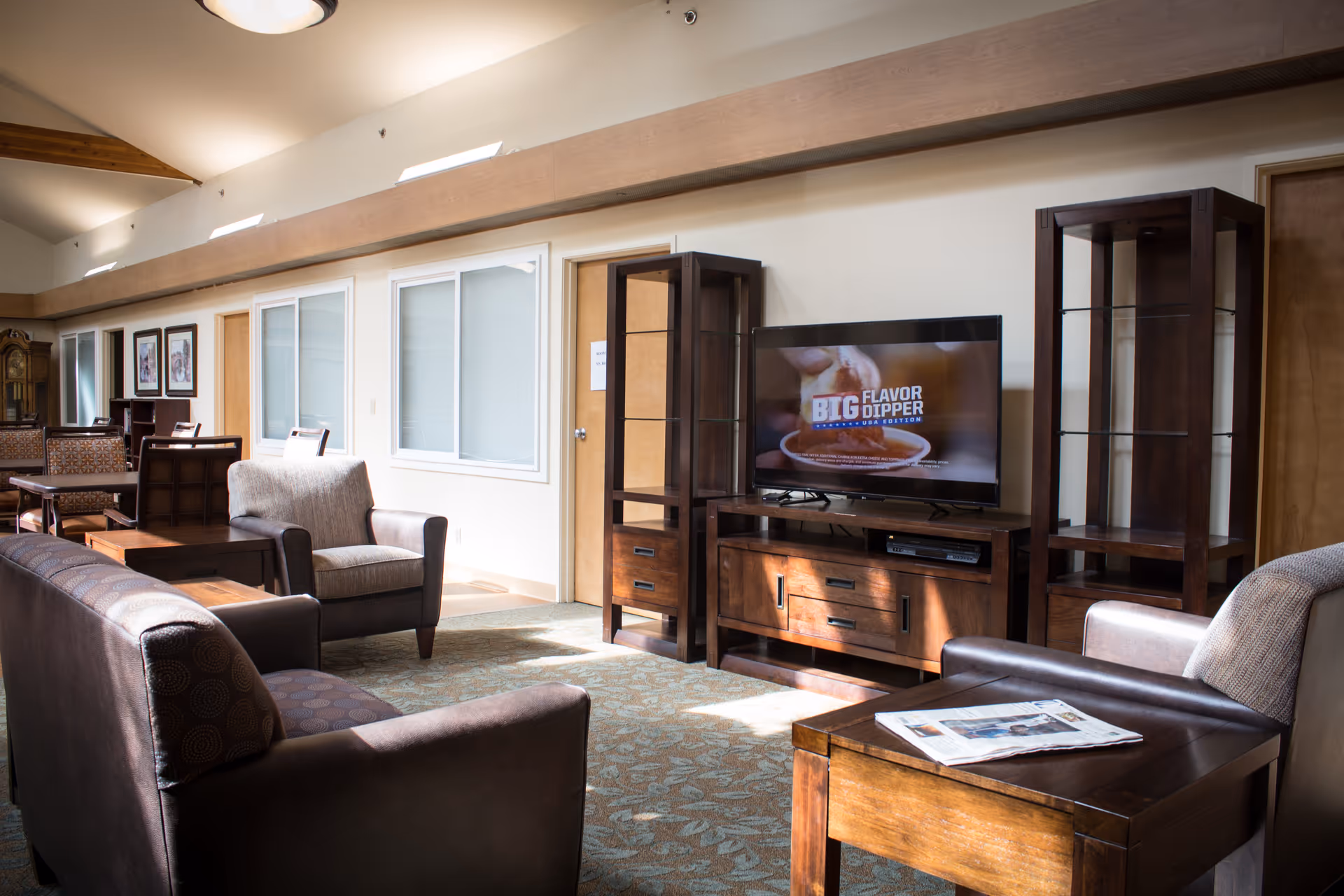 A well-lit living room area in a senior living facility featuring multiple armchairs and sofas arranged around a wooden coffee table and TV stand. A flat-screen TV displays an advertisement for Big Flavor Dipper. The room has a high ceiling with exposed wooden beams, carpeted floor, and several framed pictures on the far wall.