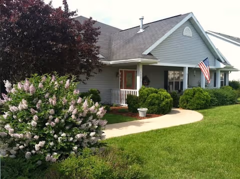 Single-story residential building with gray siding and a gable roof, featuring a small covered porch with white railing. An American flag is mounted near the porch. The front yard has green grass, bushes, a tree with dark red leaves, and a flowering shrub with light purple blossoms. A curved concrete walkway leads to the porch.