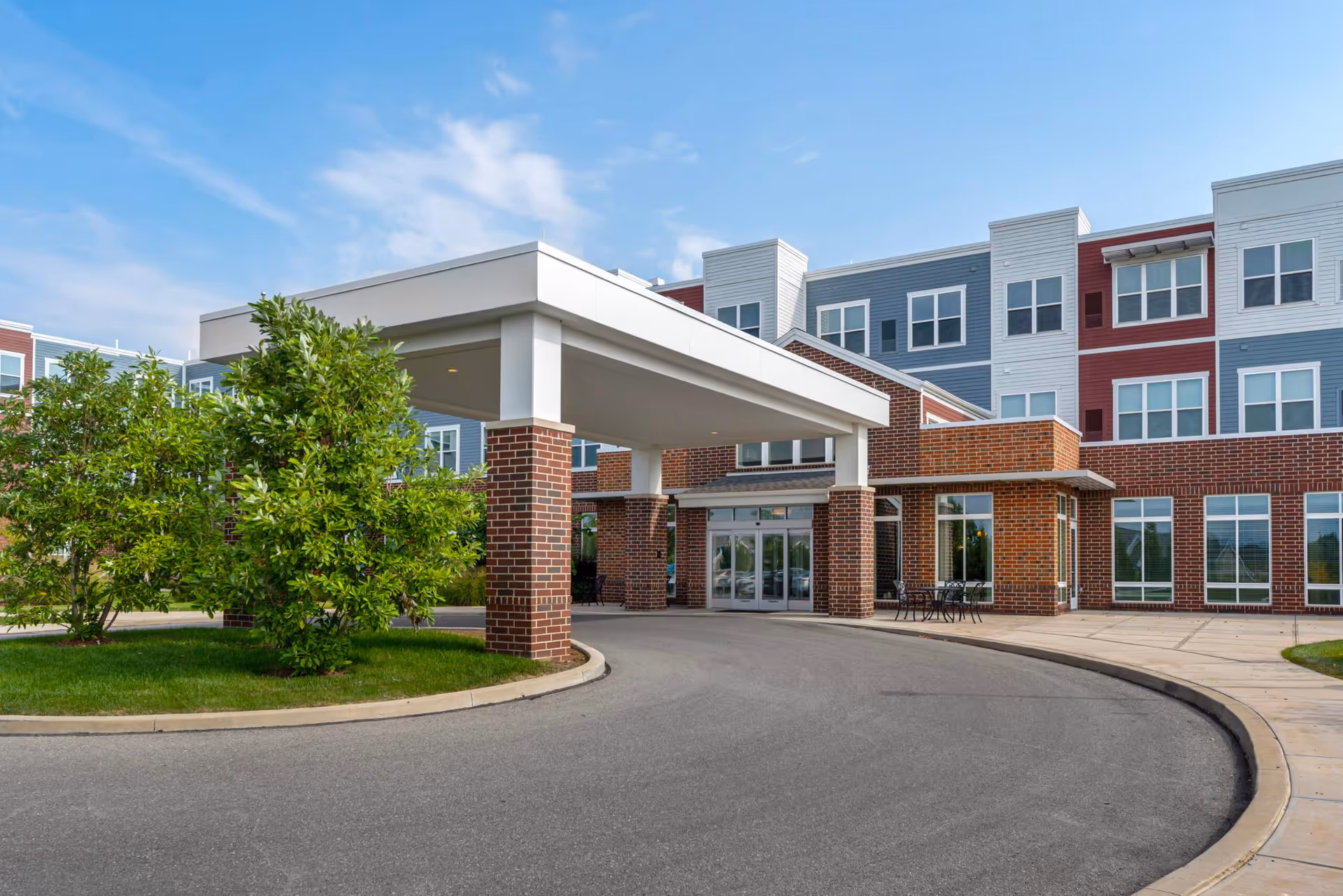 Entrance canopy and porte-cochère of a multi-story brick and siding senior living building with driveway and landscaping.