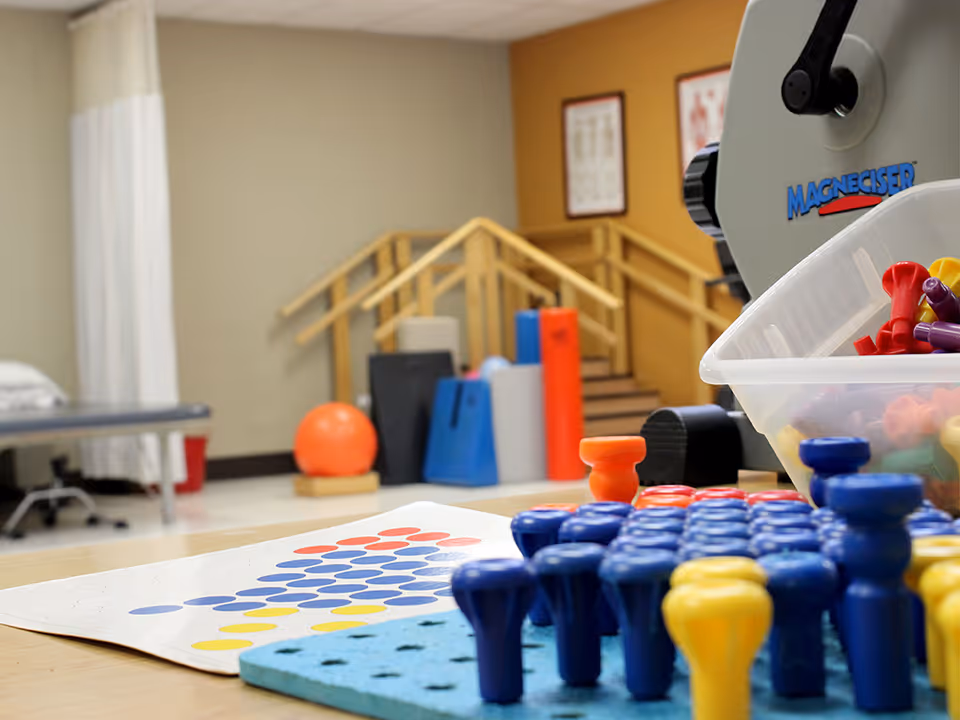 A therapy or rehabilitation room with various physical therapy equipment including a pegboard with colorful pegs, an exercise ball, foam rollers, and a set of stairs with handrails in the background.