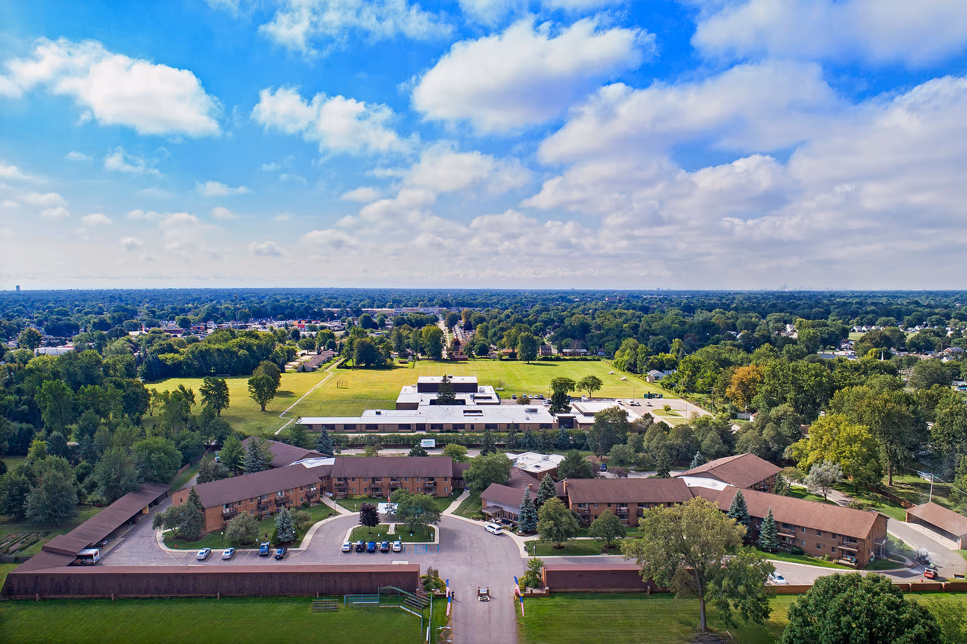 Aerial view of American House East II senior living facility surrounded by trees and greenery under a partly cloudy blue sky.