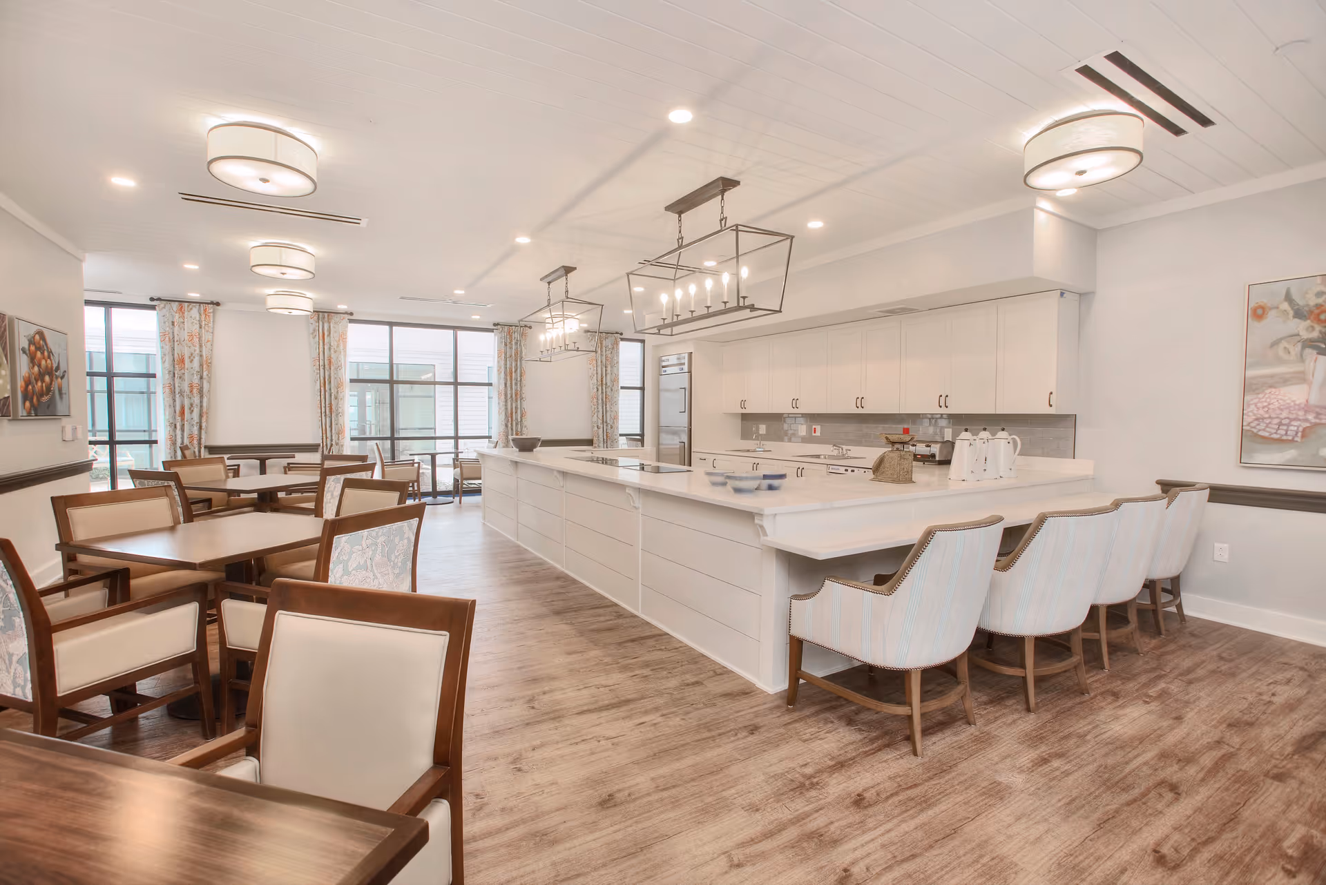 A bright and spacious dining area with multiple wooden tables and chairs on the left side and a large white kitchen island with bar stools on the right. The room features modern light fixtures, large windows with floral curtains, and light-colored walls and flooring.