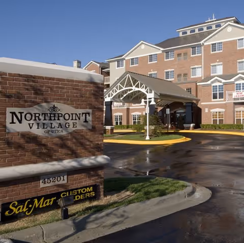 Exterior view of Northpoint Village of Utica senior living facility showing a multi-story brick building with white trim, a covered entrance, and a sign with the facility name in front.