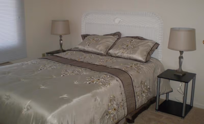 A neatly made bed with a white wicker headboard, beige and brown floral bedding, and two matching pillows. On each side of the bed is a small black nightstand with a silver lamp. A window with closed blinds is visible on the left side of the room.