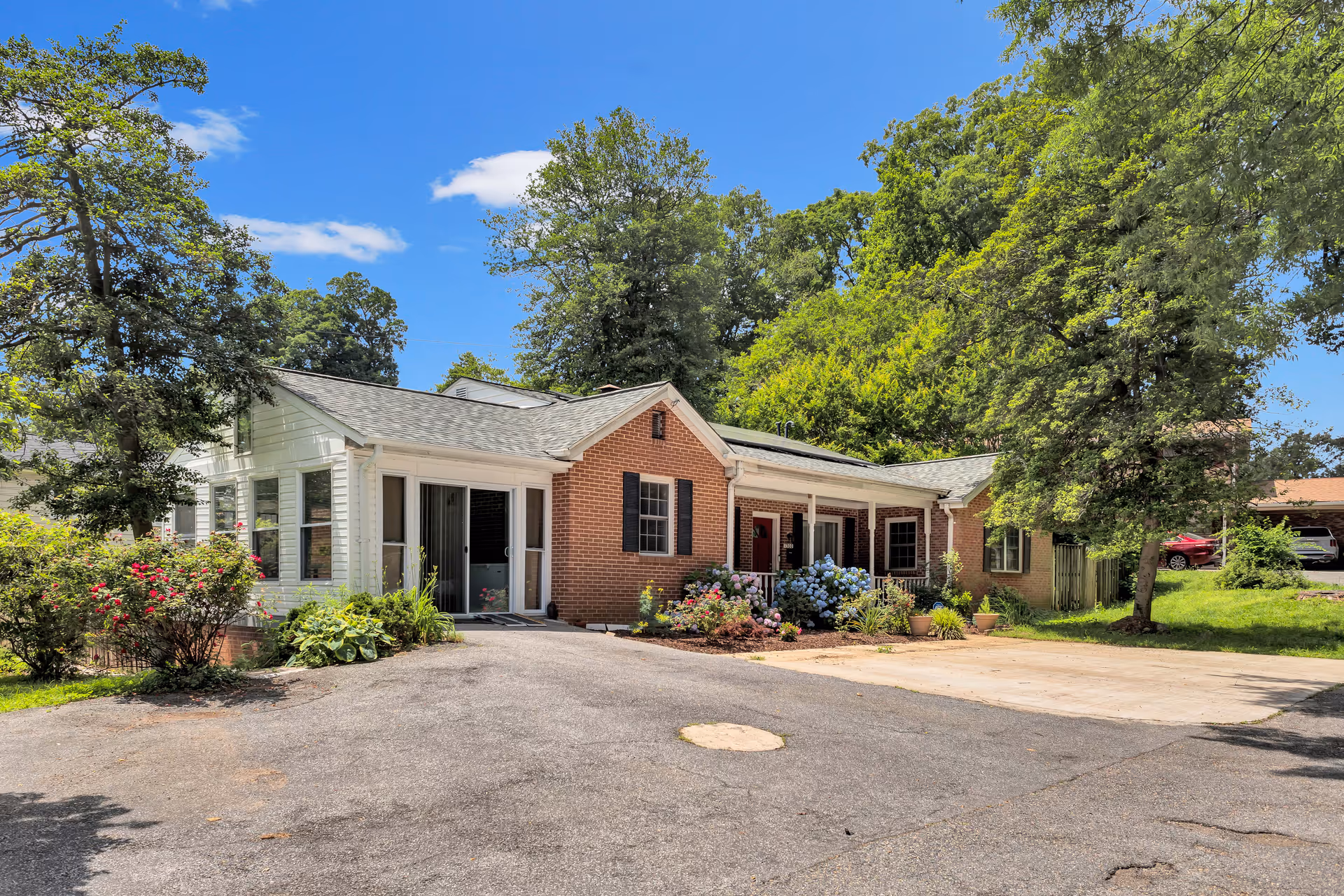 Single-story brick and white siding building with a front porch, surrounded by greenery and flowers under a clear blue sky.