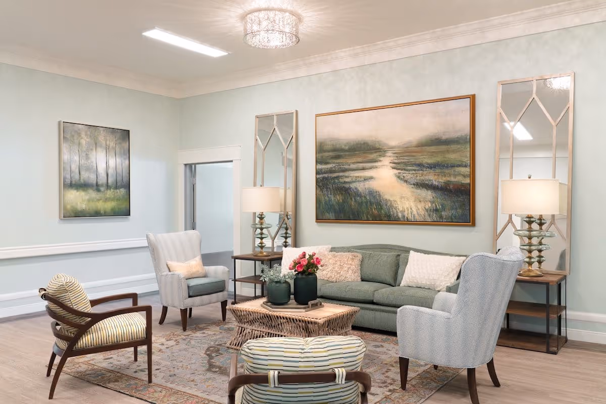 A cozy living room with a green sofa adorned with white and beige pillows, two light gray armchairs, and two wooden chairs with striped cushions. A wicker coffee table with flower vases sits on a patterned rug. The walls are light blue with two large landscape paintings and two tall mirrors flanking a pair of table lamps on side tables.