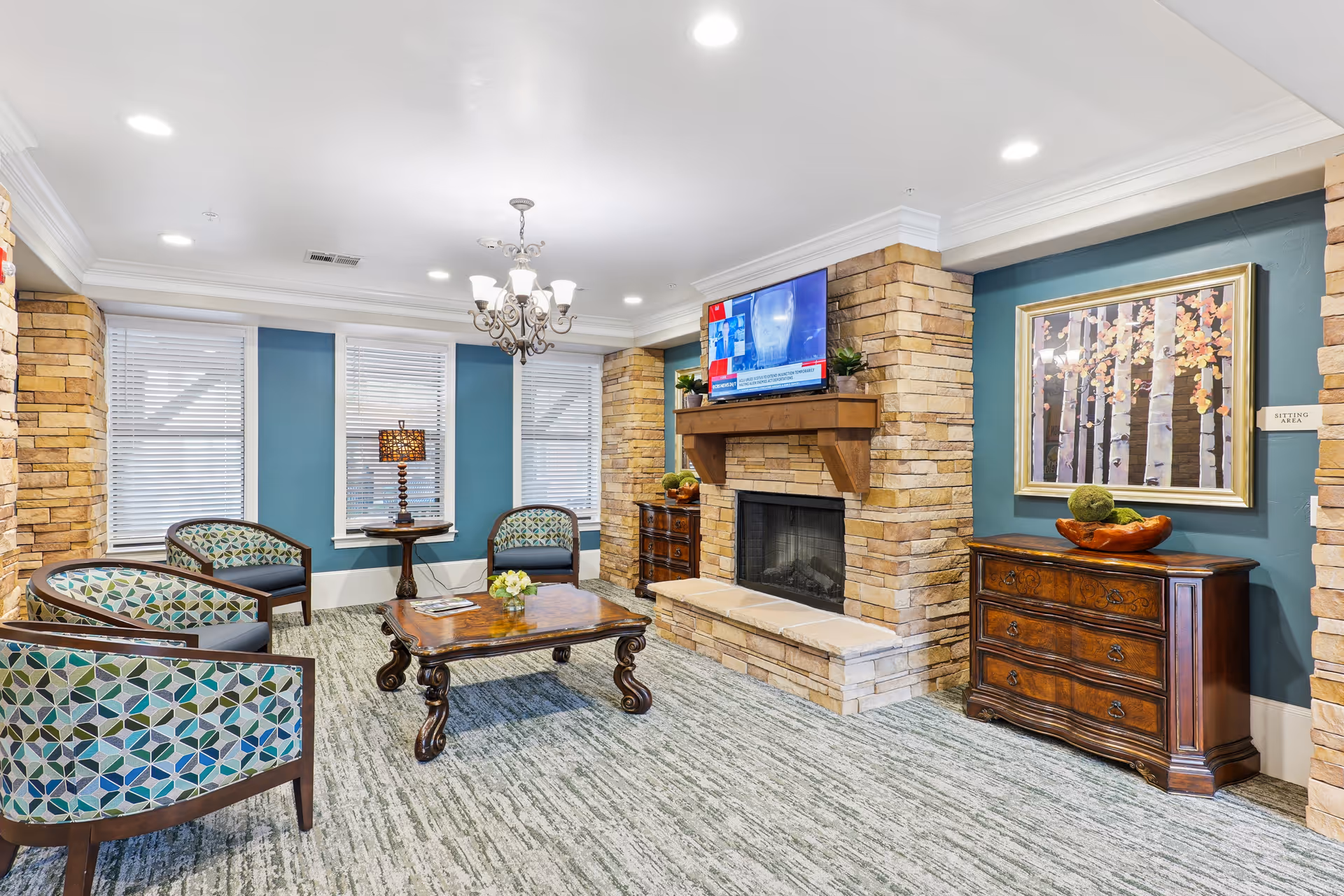 A cozy sitting area with four patterned armchairs arranged around a wooden coffee table on a carpeted floor. The room features a stone fireplace with a mounted flat-screen TV above it, blue walls with white trim, three windows with blinds, a chandelier, a table lamp on a small round table, and a wooden dresser with decorative items and a framed painting of birch trees on the wall.