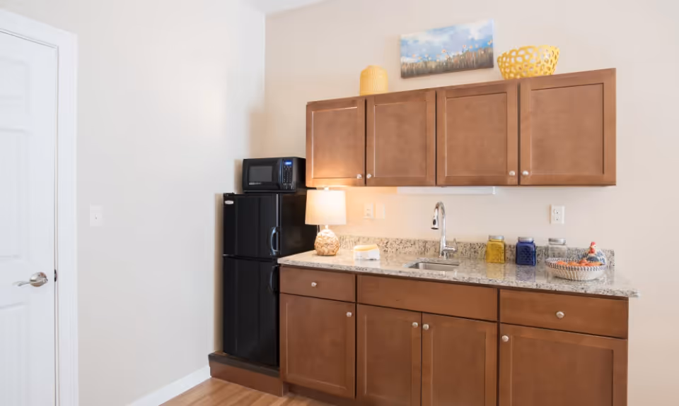 A small kitchenette area with brown wooden cabinets, a granite countertop, a stainless steel sink with a faucet, a black mini refrigerator with a microwave on top, a table lamp, decorative jars, and a basket. A painting of flowers hangs on the wall above the cabinets.