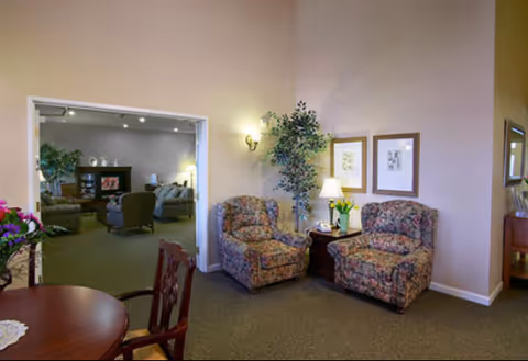 A cozy senior living common area with two floral upholstered armchairs positioned around a small wooden side table with a lamp and a vase of flowers. The walls are light beige with two framed pictures hanging above the chairs. A tall potted plant is placed in the corner. In the background, through a large open doorway, another seating area with additional chairs and a wooden cabinet is visible. A round wooden table with chairs is partially visible in the foreground.