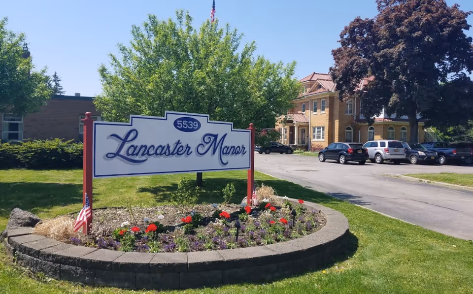 Outdoor view of Lancaster Manor facility sign surrounded by a flower bed with red and purple flowers, green grass, and trees. In the background, there is a parking lot with several cars and a large brick building with a red roof.