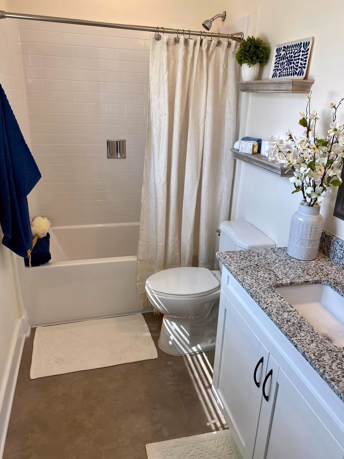 Bright bathroom with a tub/shower behind a beige curtain, a toilet, granite-top vanity, wall shelves, and decorative plants and towels.