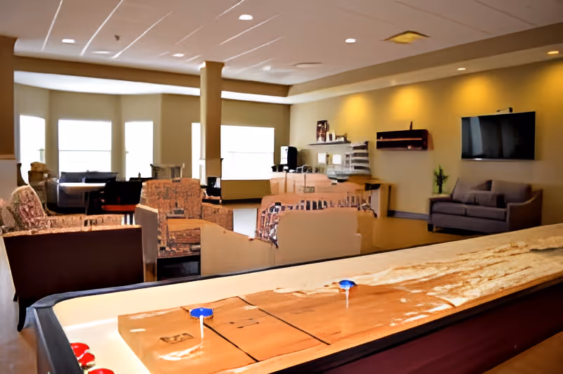 A spacious common area with multiple armchairs and sofas arranged around the room. Large windows allow natural light to fill the space. A flat-screen TV is mounted on the wall above a small couch. In the foreground, there is a shuffleboard table with pucks on it. The room has a warm and inviting atmosphere with beige walls and ceiling lights.