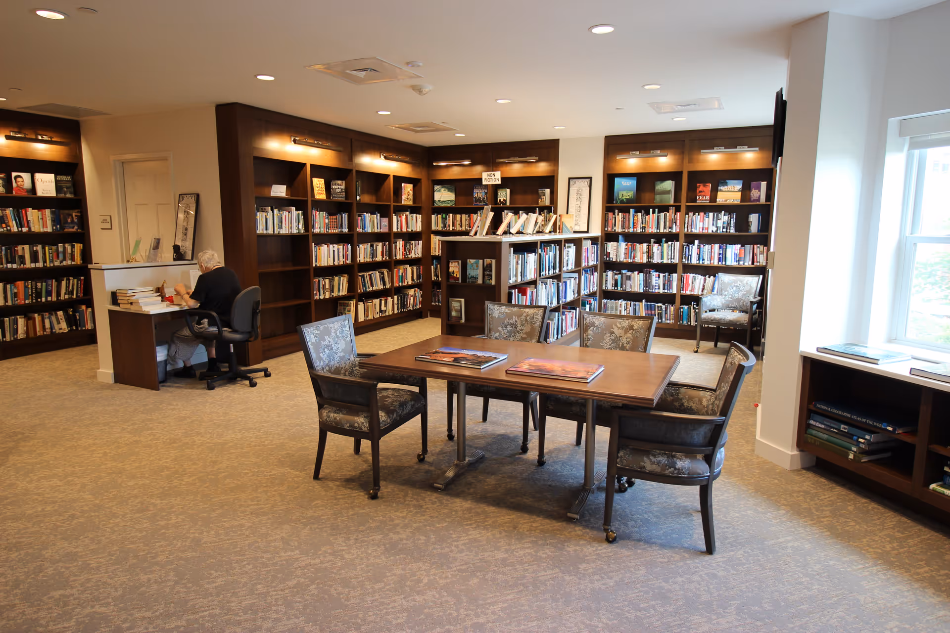 A well-lit library room with wooden bookshelves filled with books along the walls and a central bookshelf. There is a wooden table with four upholstered chairs around it, and two large books are placed on the table. An elderly person is seated at a small desk in the corner, reading or writing. A window on the right side lets in natural light.