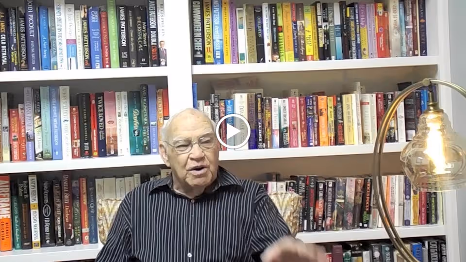 An elderly man wearing glasses and a striped shirt is sitting in front of a large bookshelf filled with books. To the right, there is a modern lamp with a visible light bulb.