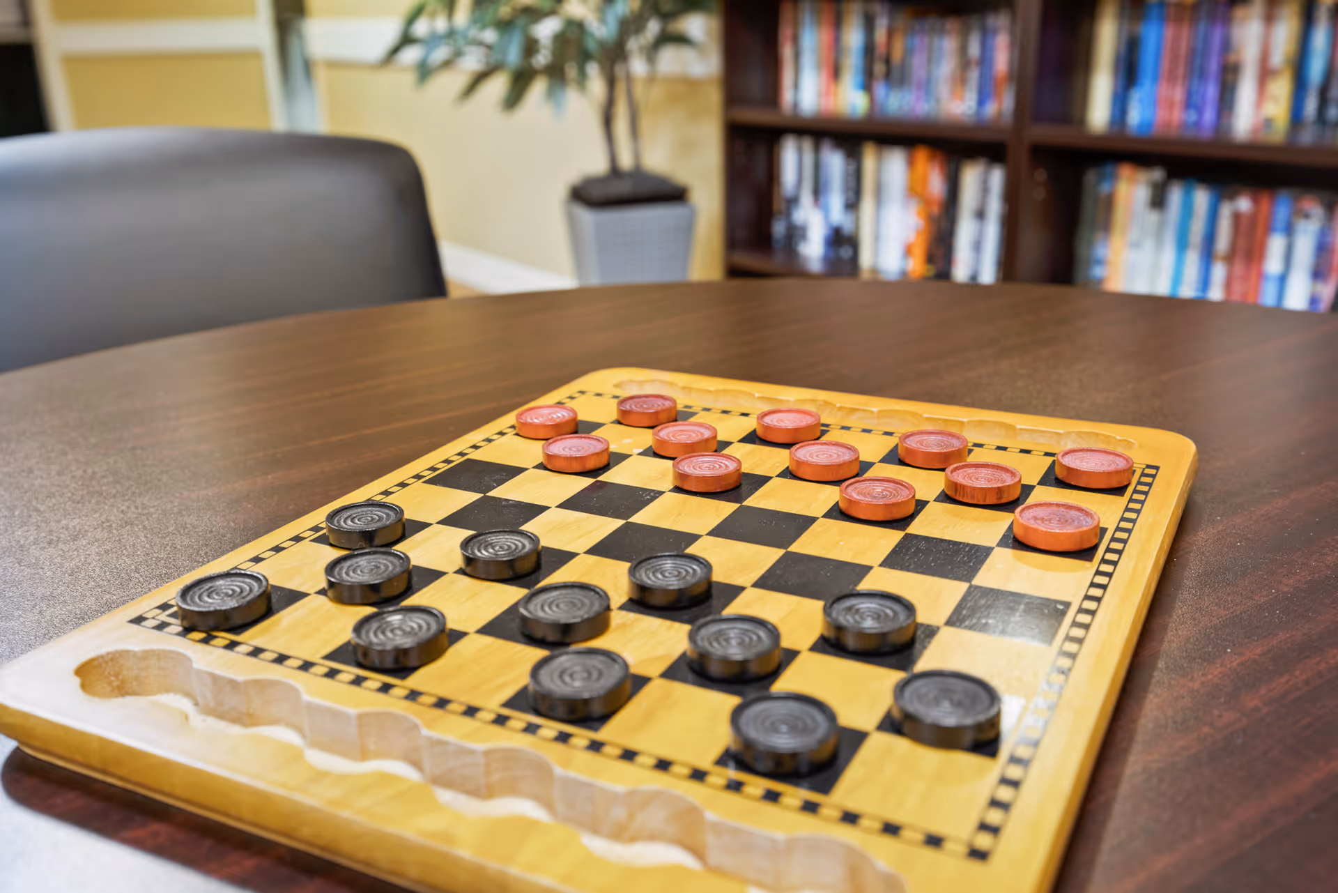 A wooden checkers board with black and red checkers pieces set up on a dark brown table. In the background, there is a black chair, a potted plant, and a bookshelf filled with books.