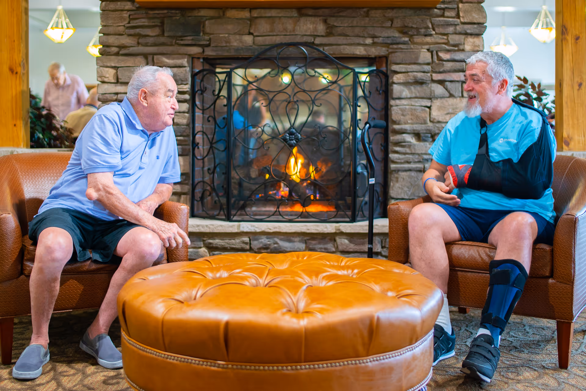 Two elderly men sitting in armchairs facing each other in a cozy room with a stone fireplace and a fire burning. One man is wearing a light blue polo shirt and black shorts, while the other man has a beard, is wearing a blue t-shirt, black shorts, and has his arm in a sling and leg in a brace. There is a large round leather ottoman in front of them and warm lighting in the background.