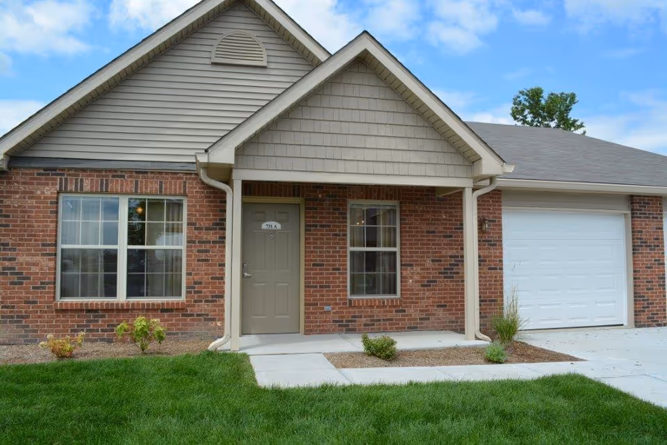 Exterior view of a single-story brick building with a beige front door labeled 711 A, two windows, a covered porch, and an attached white garage door. There is a small landscaped area with grass and plants in front of the building under a partly cloudy sky.