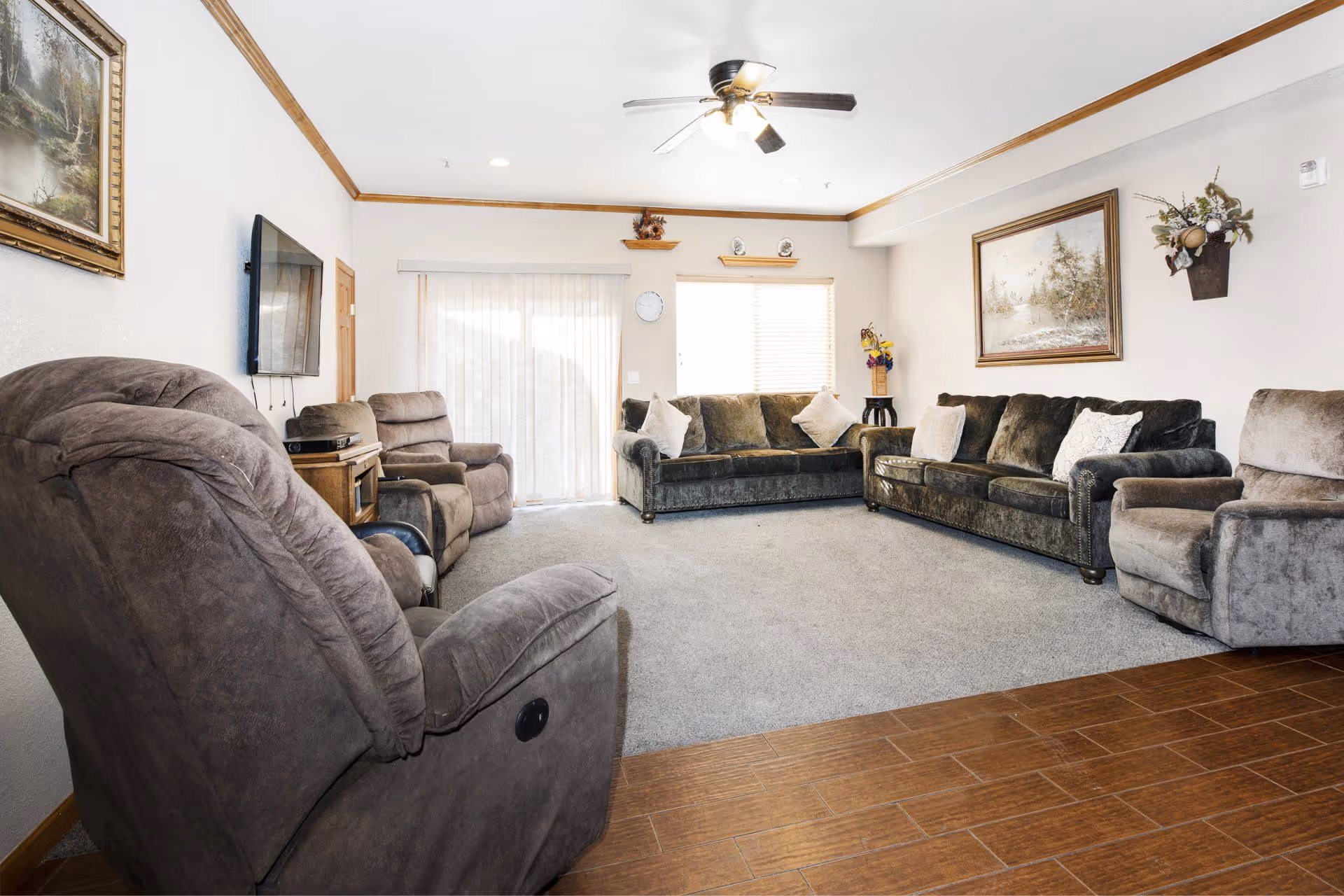 A spacious living room with multiple plush gray recliners and sofas arranged around the perimeter. The room features a ceiling fan with lights, a wall-mounted flat-screen TV, a wooden side table, and framed artwork on the walls. There is a large window with blinds and a sliding glass door with sheer curtains allowing natural light to enter. The floor is a combination of wood and carpet.