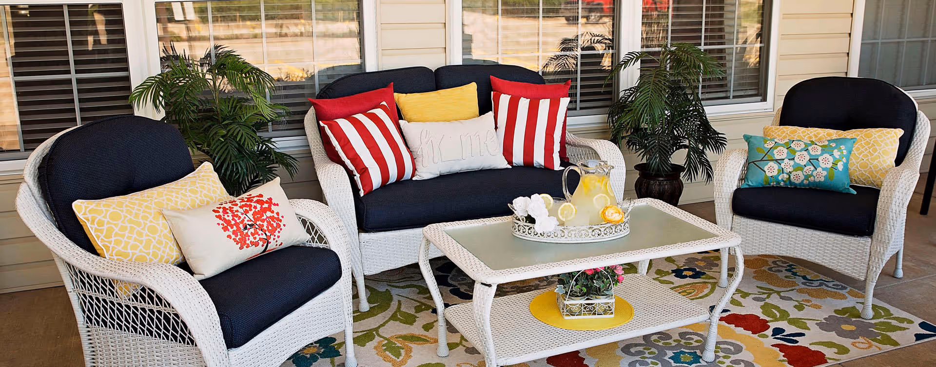 A cozy outdoor seating area with white wicker furniture including two armchairs and a loveseat with dark blue cushions and colorful decorative pillows. A white coffee table with a glass pitcher of lemonade and glasses sits on a floral patterned rug. There are green potted plants behind the loveseat and armchairs, and windows with blinds in the background.