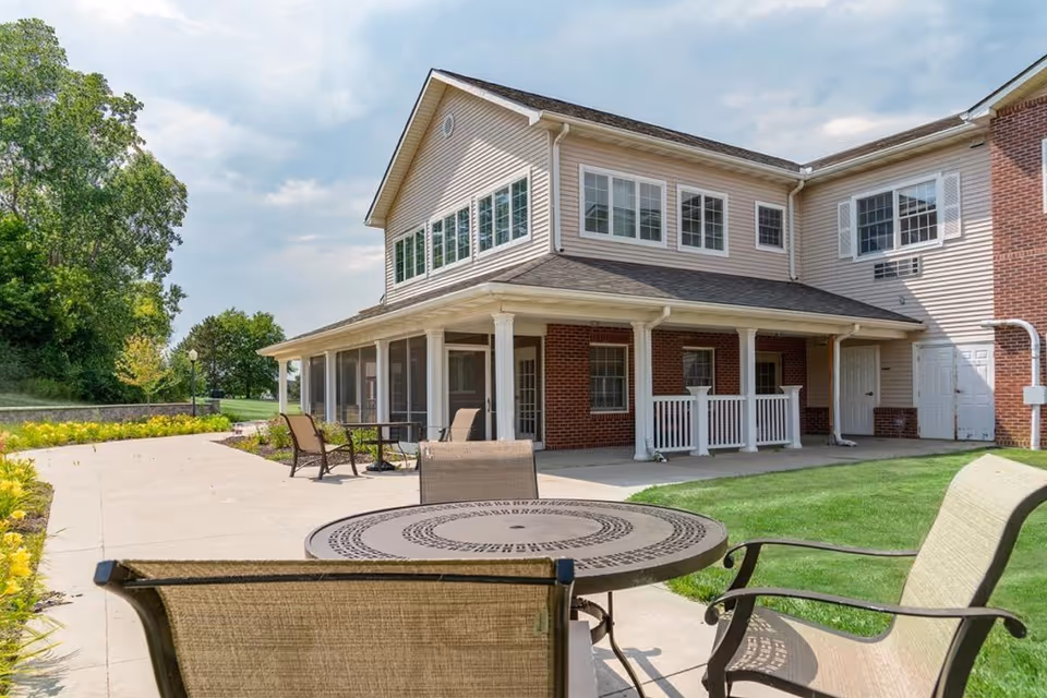 Outdoor patio area at Landings of Genesee Valley with metal table and chairs on a concrete walkway, adjacent to a two-story building with brick and beige siding, surrounded by green grass and trees under a partly cloudy sky.