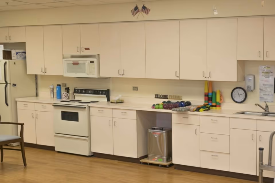 A kitchen area with white cabinets, a white stove, microwave, and refrigerator. Various items are on the countertop including water bottles, a tissue box, colorful exercise weights, and stacked plastic cups. A clock and a hand sanitizer dispenser are mounted on the wall above the sink. Two small American flags are displayed above the cabinets.