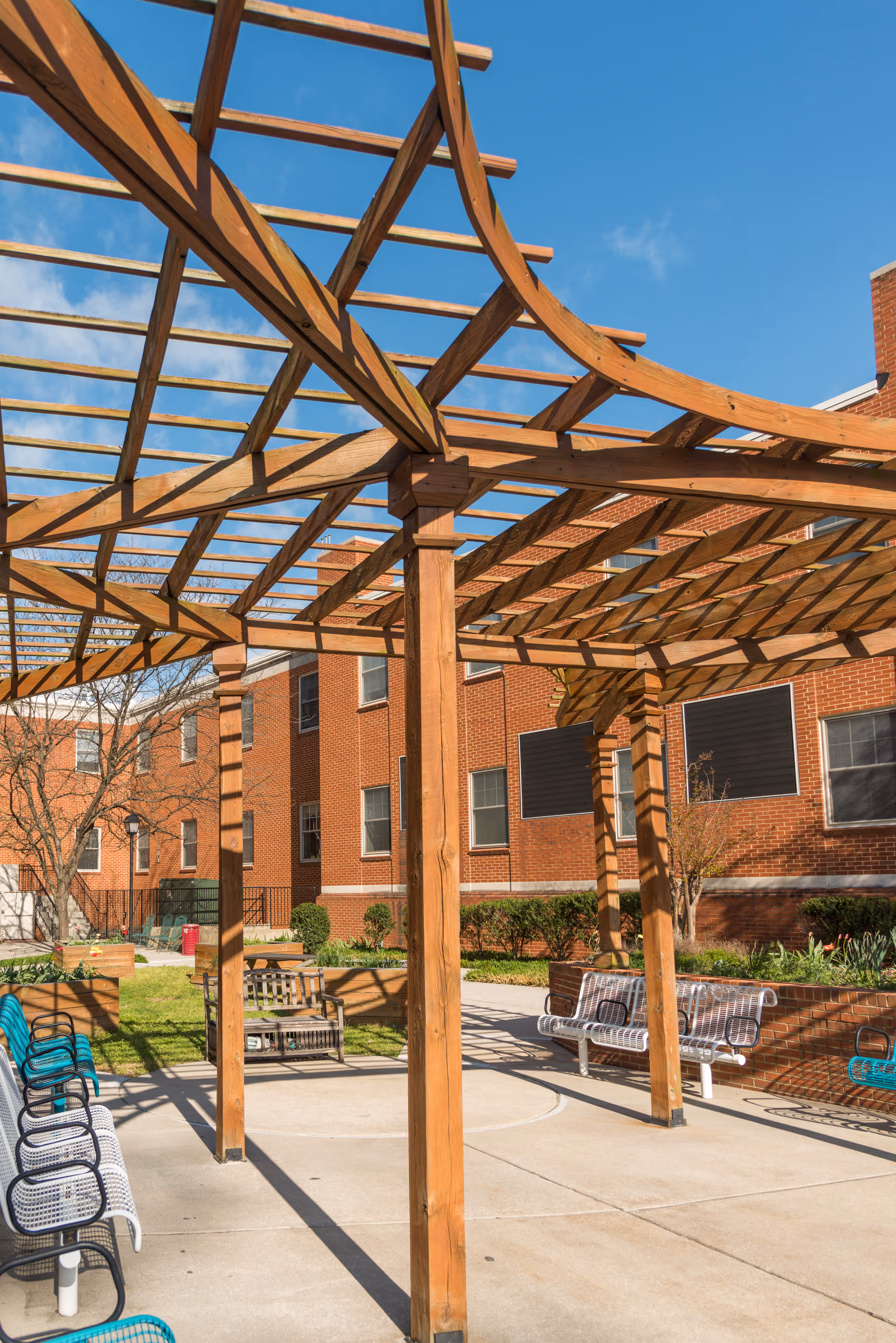 Outdoor seating area with wooden pergola structure casting shadows on the concrete ground, surrounded by benches and chairs, with a brick building and garden beds in the background under a clear blue sky.