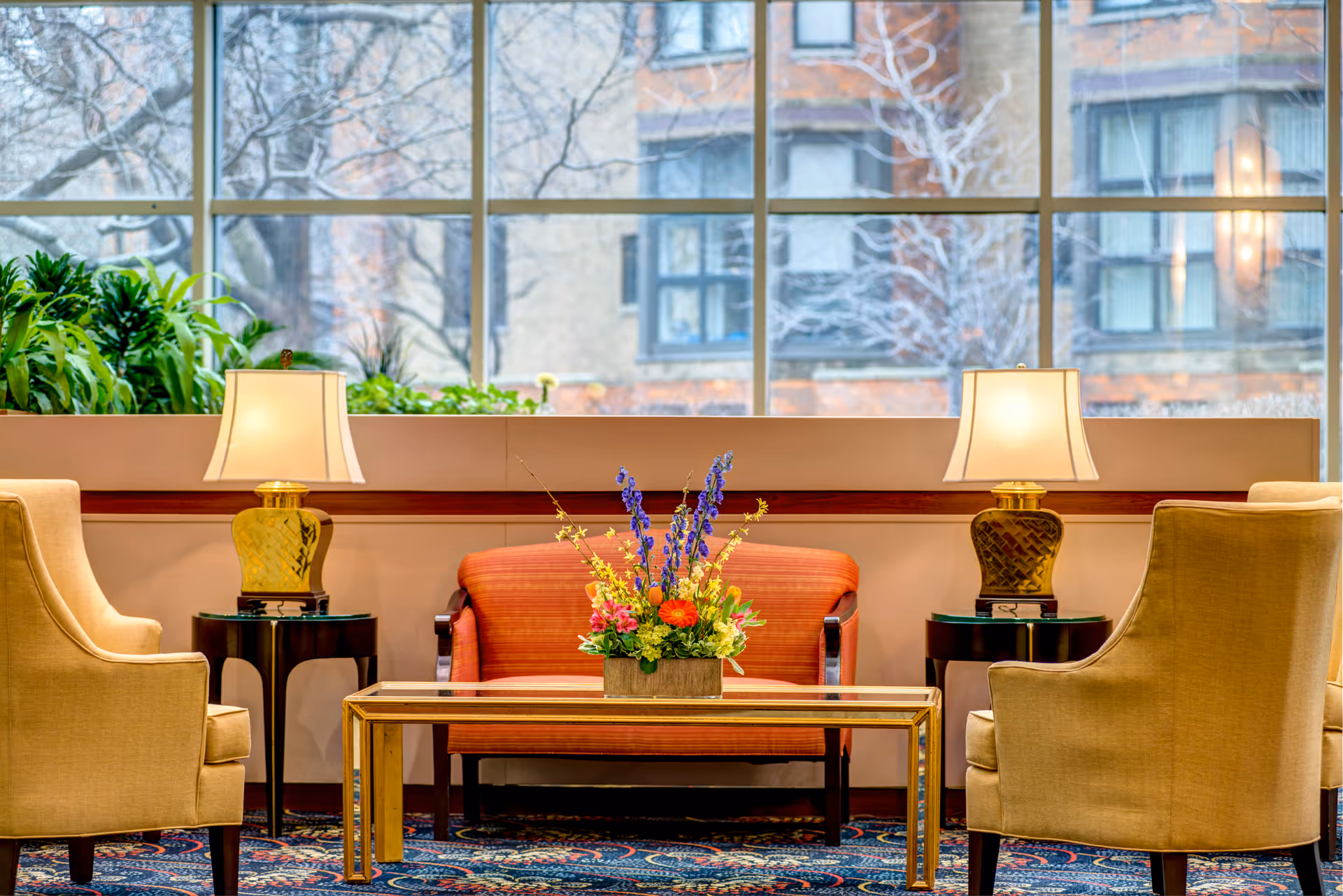 A cozy sitting area with a red loveseat, two beige armchairs, a glass coffee table with a colorful flower arrangement, two table lamps on dark side tables, and large windows showing an outside view of buildings and trees.