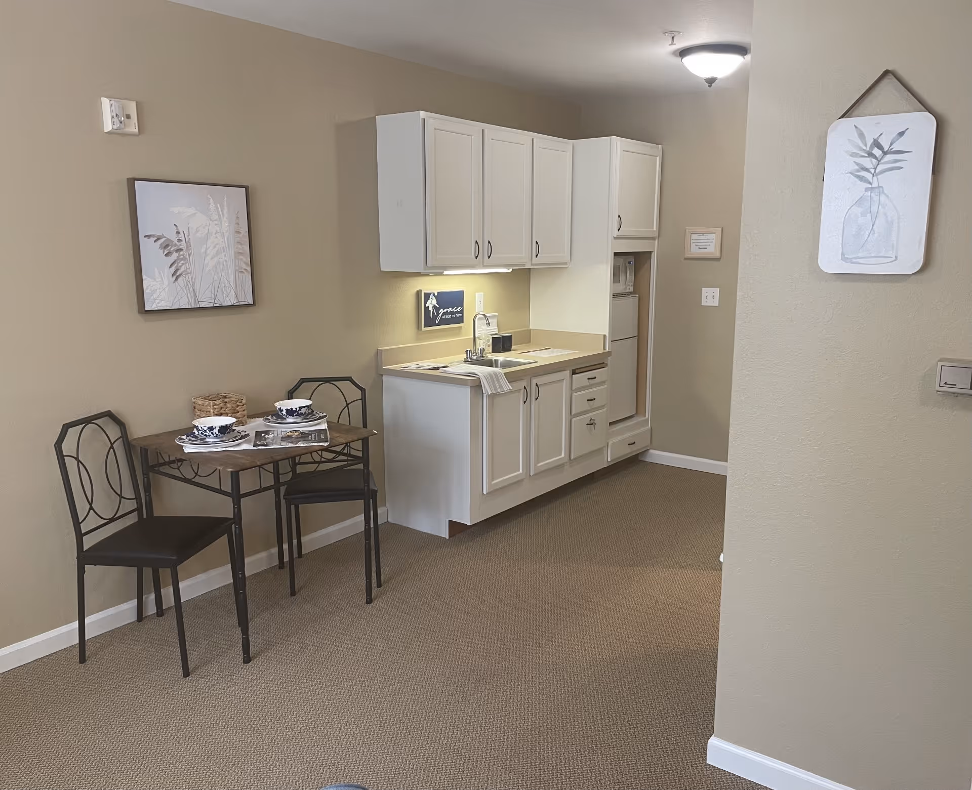 A small kitchenette area with white cabinets, a sink, and a compact refrigerator. Next to the kitchenette is a small dining table set with two chairs, two bowls, and a basket. The walls are beige with two framed botanical prints hanging on them. The floor is carpeted in a neutral tone.