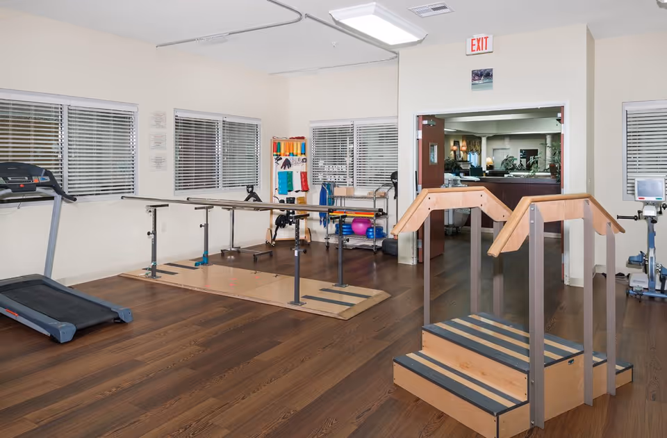 A rehabilitation or physical therapy room with wooden parallel bars for walking exercises, a treadmill, exercise equipment, and a small set of wooden stairs with handrails. The room has large windows with blinds, wood flooring, and an exit door leading to another area.