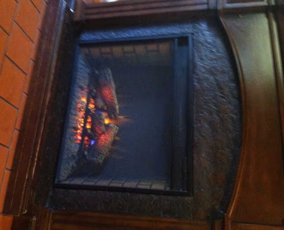 A close-up view of a lit indoor fireplace with glowing embers and logs behind a protective mesh screen, surrounded by a dark textured frame and wooden paneling.