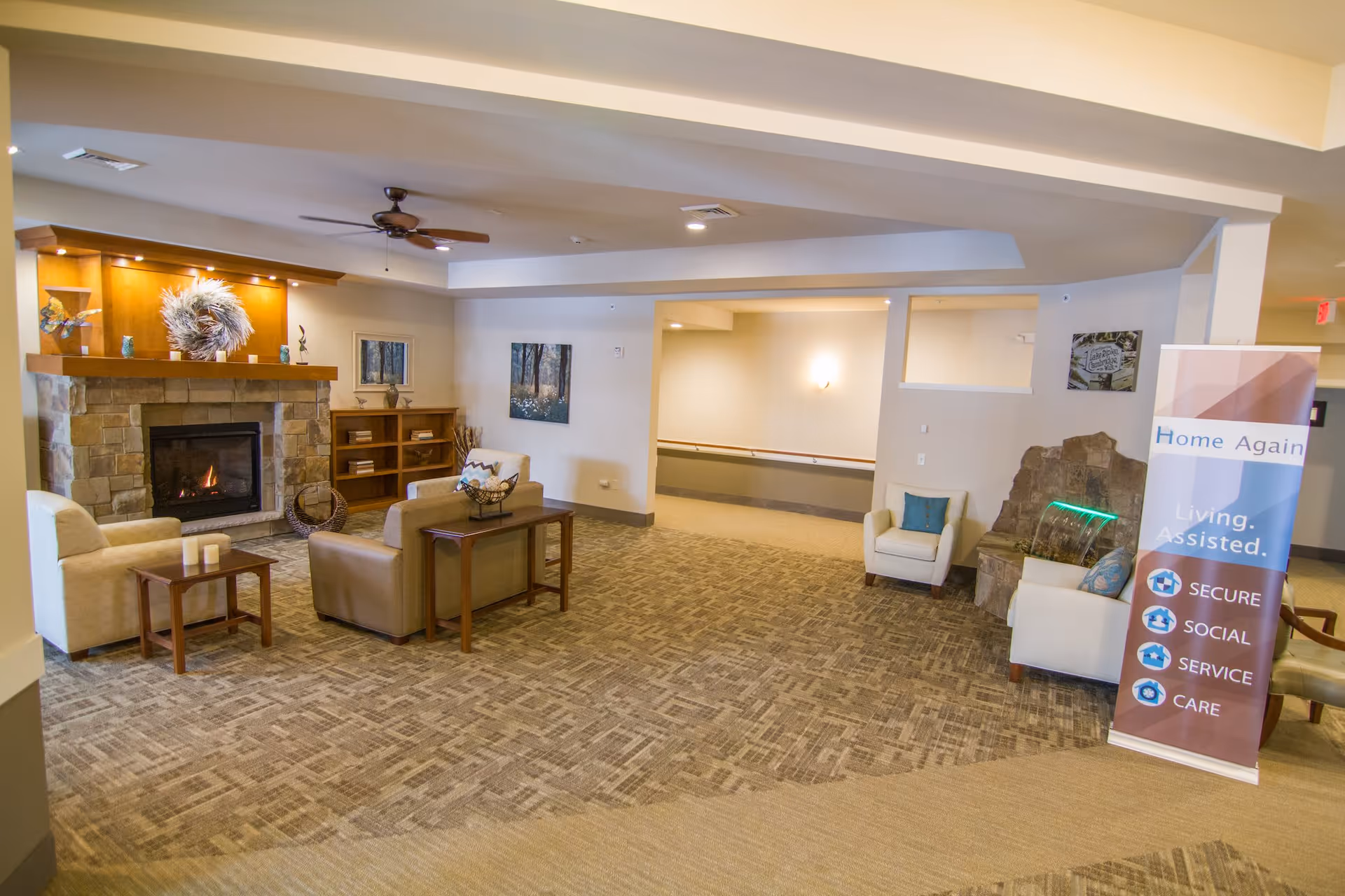 A cozy assisted living facility common area with a stone fireplace, beige armchairs, a sofa, wooden side tables, and a ceiling fan. There is a banner on the right side that reads 'Home Again Living. Assisted. Secure, Social, Service, Care.' The room has carpeted flooring and soft lighting.