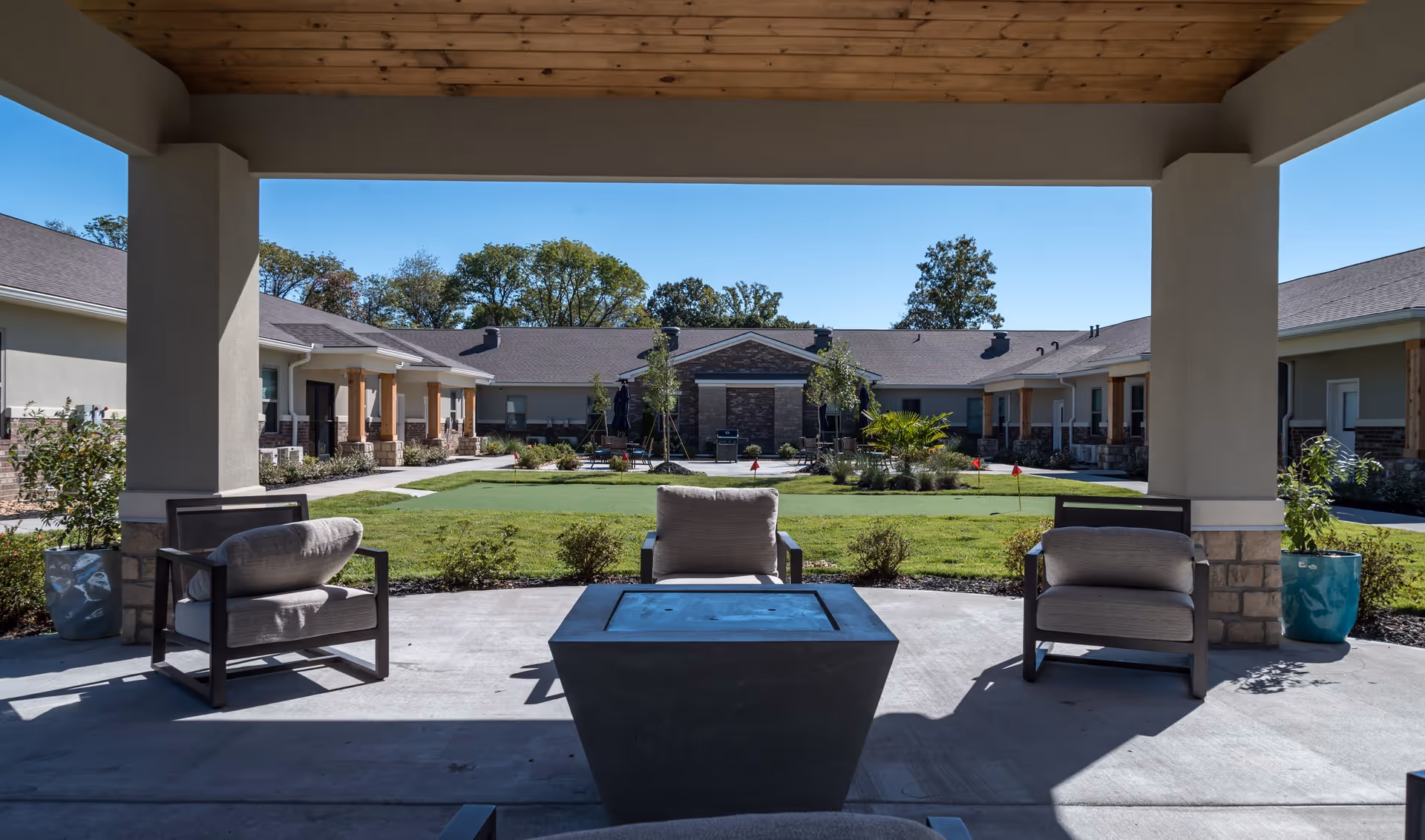 Covered outdoor seating area with cushioned chairs and a fire pit table, overlooking a green lawn with small trees and a putting green, surrounded by a single-story building under a clear blue sky.