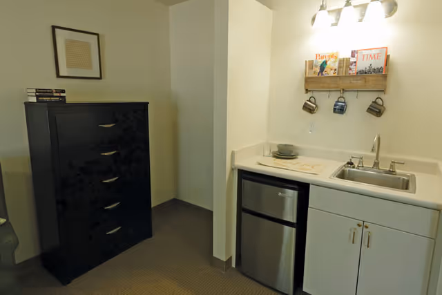 A small kitchenette area with a stainless steel mini refrigerator, a sink, and white cabinets. Above the sink is a wooden rack holding three coffee mugs and some magazines. To the left of the kitchenette is a black chest of drawers with a few books on top and a framed picture hanging on the wall above it.