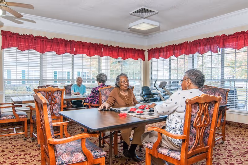 Four elderly women sitting around two tables in a bright room with large windows and red valance curtains. Two women in the foreground are playing checkers, while the other two women in the background are engaged in conversation. The room has floral upholstered wooden chairs and a patterned carpet.