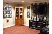 Interior view of a living room area with beige walls and carpeted floor. There is a wooden corner cabinet with glass doors displaying items inside, and a black entertainment center holding a large flat-screen TV and framed photos. Several framed pictures are hung on the walls around the room.