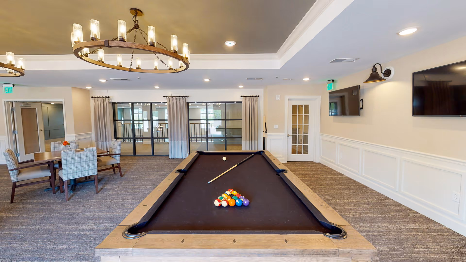 A well-lit recreational room featuring a pool table with a full set of billiard balls and a cue stick arranged on it. To the left, there is a dining table with six upholstered chairs and a small flower vase. The room has beige walls with white wainscoting, two large flat-screen TVs mounted on the right wall, and large windows with curtains at the back. Two circular chandeliers hang from the ceiling, and there are exit signs above the doors.