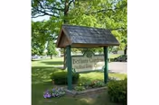 A wooden sign for Bethany Gardens Skilled Living Center mounted on two posts with a shingled roof above it, surrounded by green grass, trees, and some small flower beds.