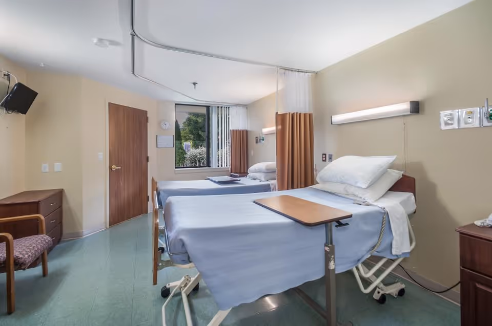 A senior facility patient room with two hospital beds, blue linens, overbed tables, privacy curtains and a window.