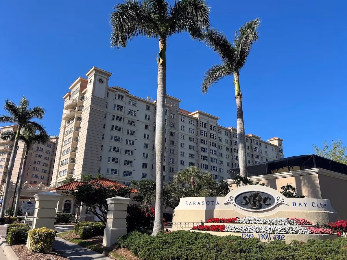 Exterior view of a large multi-story residential building with palm trees and a clear blue sky. In front of the building is a landscaped area with a sign that reads 'Sarasota Bay Club' surrounded by colorful flowers.