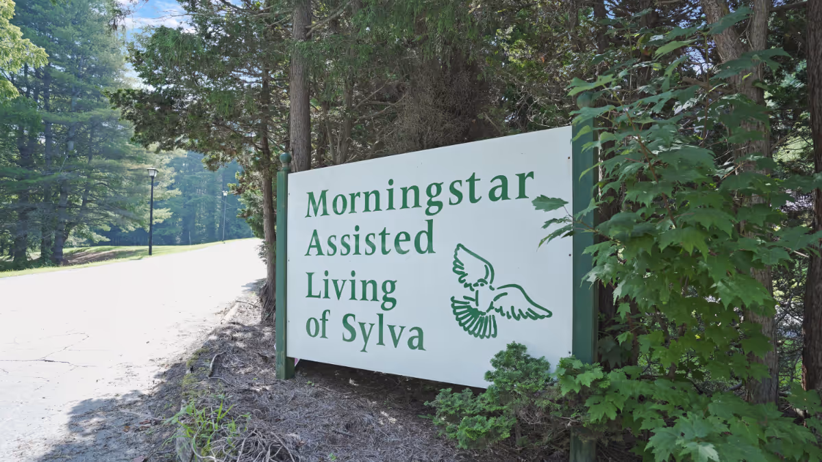A white exterior sign with green lettering reading 'Morningstar Assisted Living of Sylva' sits beside a tree-lined road.