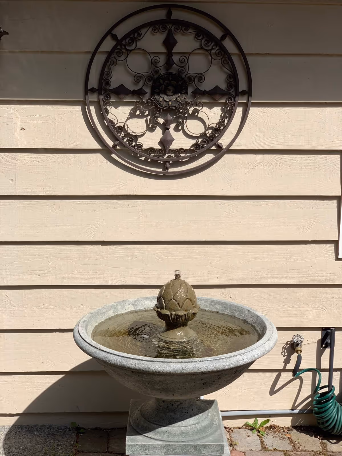 A stone water fountain with a decorative finial in the center, placed against a beige wooden exterior wall. Above the fountain is a round, ornate metal wall decoration. To the right of the fountain, a green garden hose is coiled and attached to an outdoor faucet.