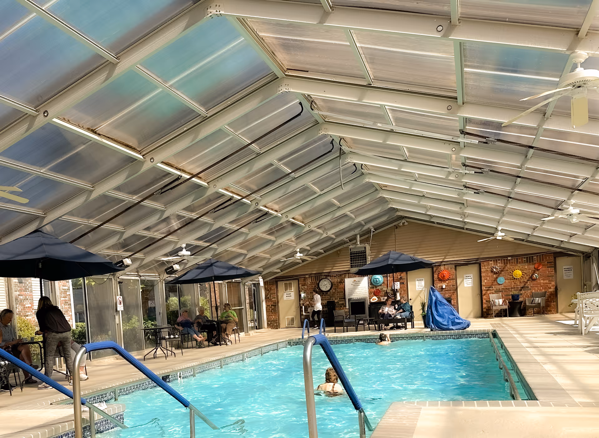 Indoor swimming pool area with a translucent pitched roof, pool ladder, umbrellas and residents seated around tables.