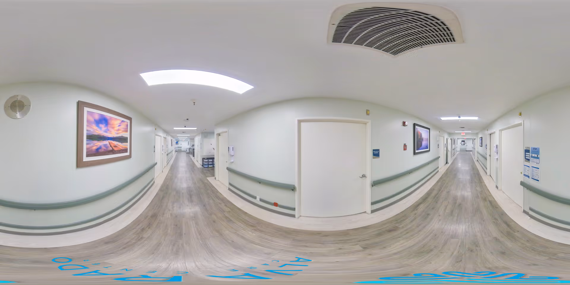 A wide, well-lit hallway in a care facility with light green walls, wooden flooring, handrails on both sides, and several closed white doors. The hallway is decorated with framed landscape pictures and has ceiling lights and ventilation. The corridor extends into the distance with a clean and orderly appearance.