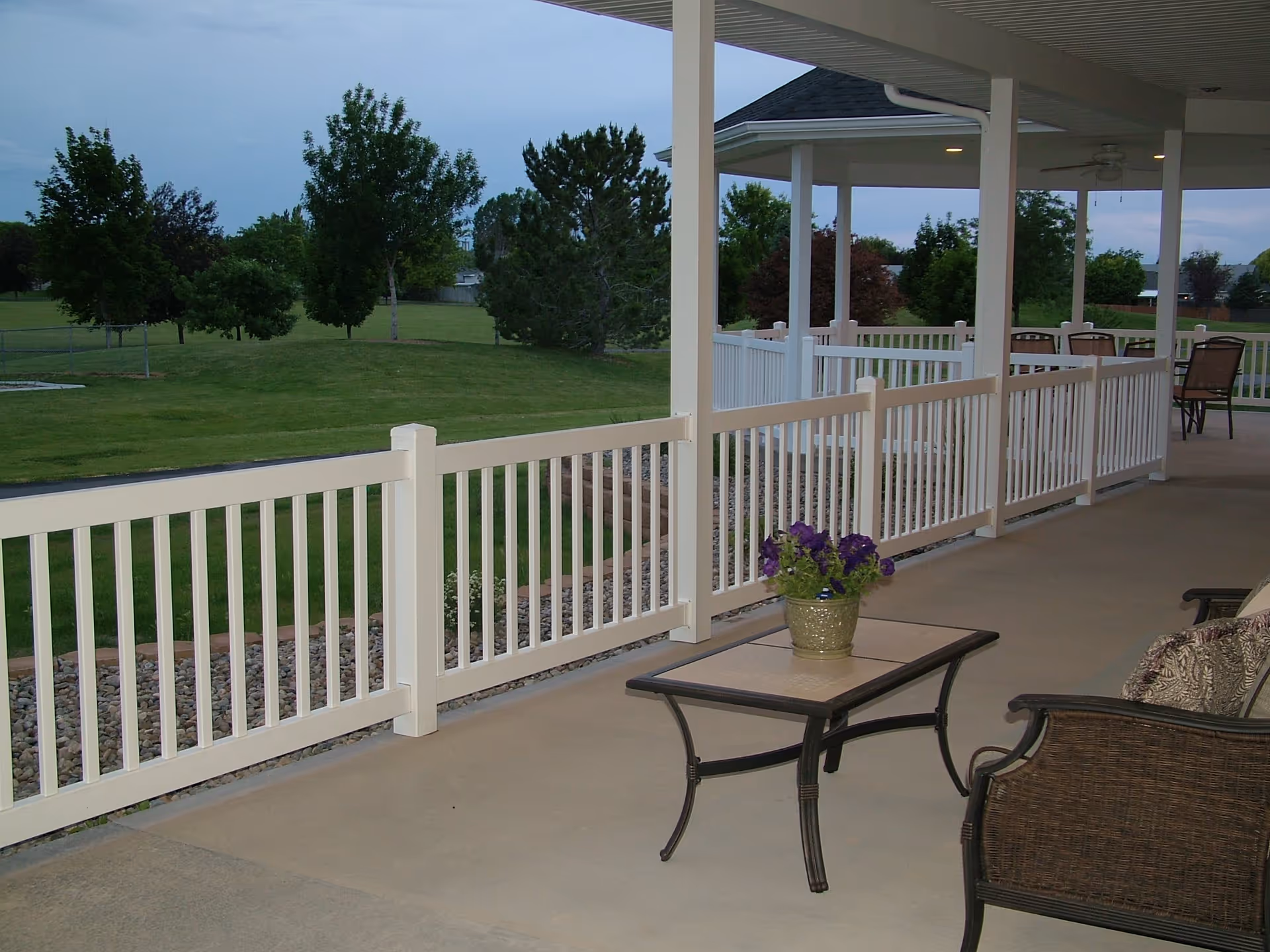Covered outdoor patio area with white railing, a small table with a potted plant with purple flowers, and wicker chairs with cushions. The patio overlooks a grassy area with trees and shrubs under a cloudy sky.