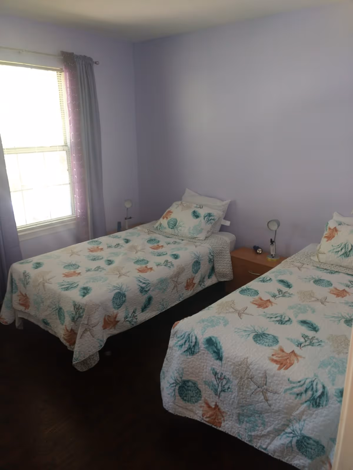 A bedroom with two twin beds covered in white bedspreads featuring a sea-themed pattern with starfish, shells, and coral. There is a wooden nightstand between the beds with two small lamps and a flashlight. A window with blinds and purple curtains is on the left wall, and the walls are painted light purple.
