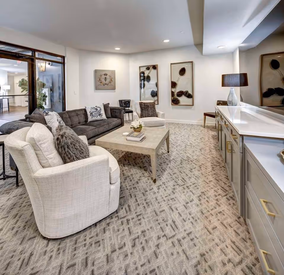 A modern living room with a gray sofa, two white armchairs with cushions, a square coffee table with books and a small plant, patterned carpet, wall art, and a sideboard with a lamp. The room has recessed lighting and a glass door leading to another area.